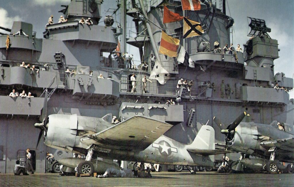 The U.S. Navy aircraft carrier USS Yorktown (CV-10) during the Marcus Island raid on 31 August 1943: Lt. Comdr. James H. "Jimmy" Flatley,Commander of Air Group 5 (CAG-5), sits in his Grumman F6F-3 Hellcat (code "00"), painted in a tricolor-scheme (certainly an "in the field" application) before takeoff. An Aviation Boatswain Mate stands ready to remove chock from wheels. A non-specular insignia white diagonal stripe on the tail and the green propeller hub signified CAG-5 aboard the Yorktown.